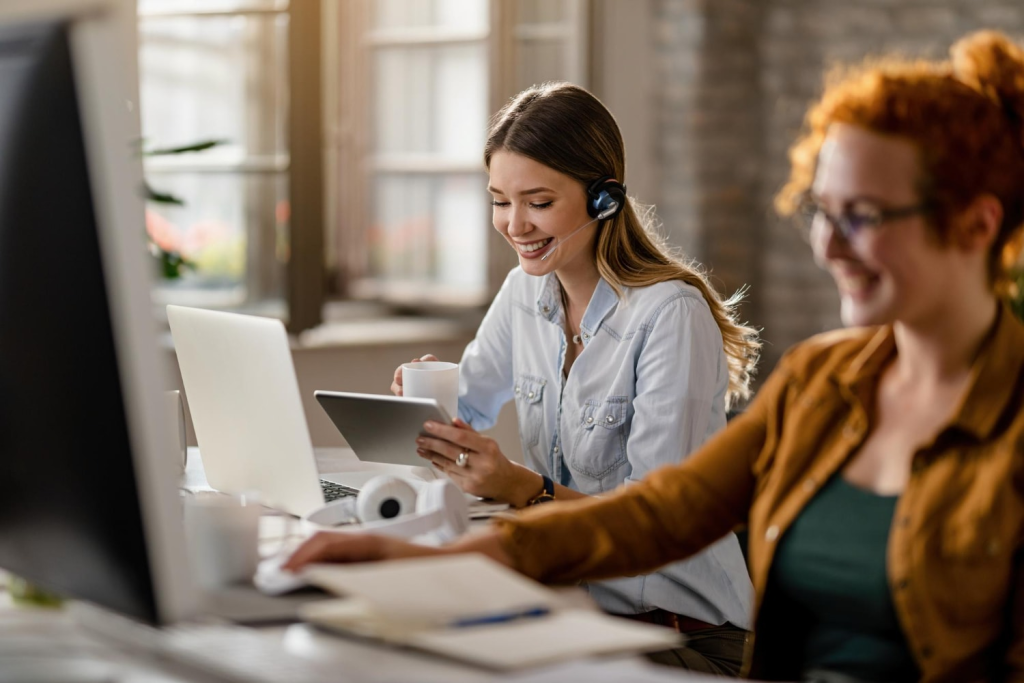 A young woman wearing a headset sits at her desk in a well-lit office, smiling as she engages with a customer through an omnichannel customer support platform. She holds a tablet while sipping coffee, surrounded by scalable contact center platforms that enhance her workflow.