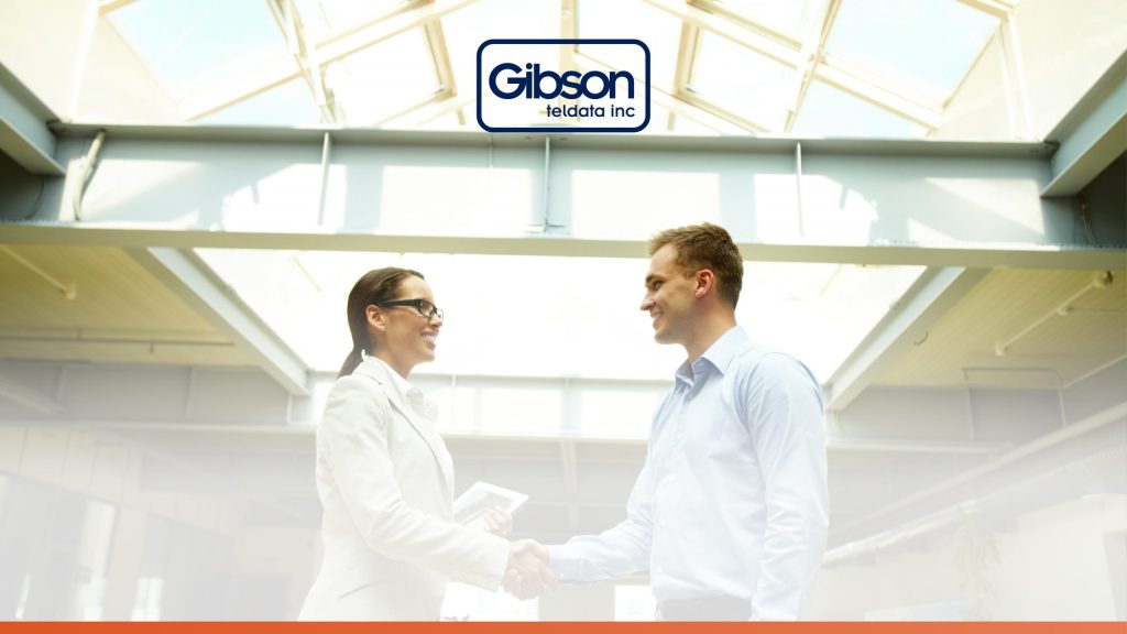 A man and woman in business attire shake hands in a bright, skylit office with the Gibson Teldata Inc. logo above. This image represents building partnerships through effective business communication solutions.