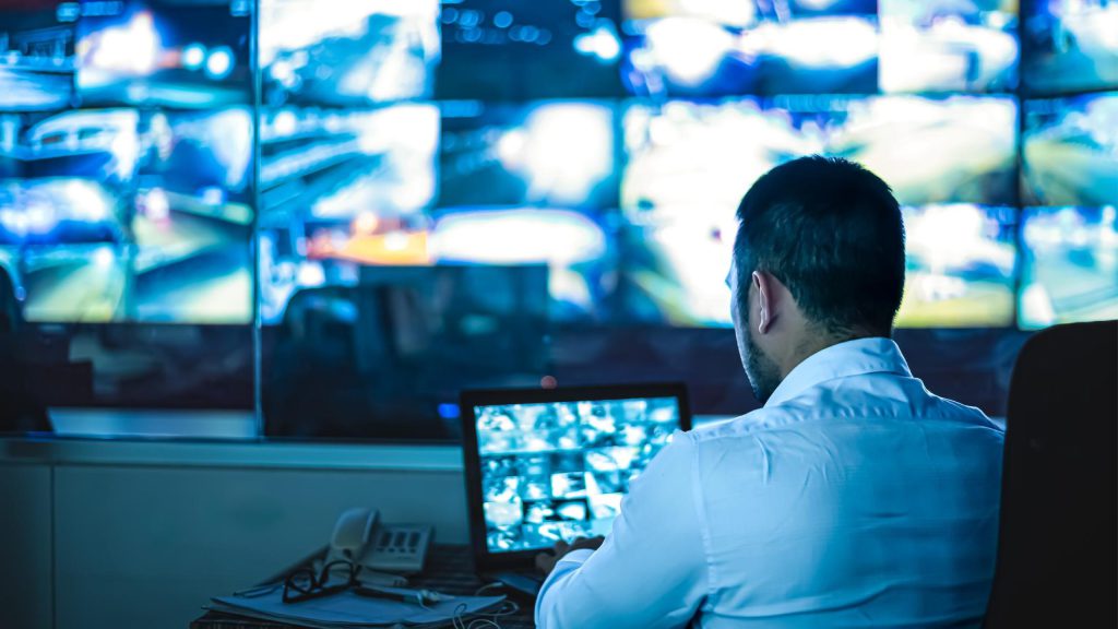 Man monitoring multiple surveillance screens in a control room, demonstrating Integrated Security Systems for real-time video monitoring and threat detection.